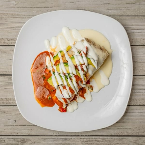 Burrito topped with red sauce, white cream, and sliced avocado on a white plate.