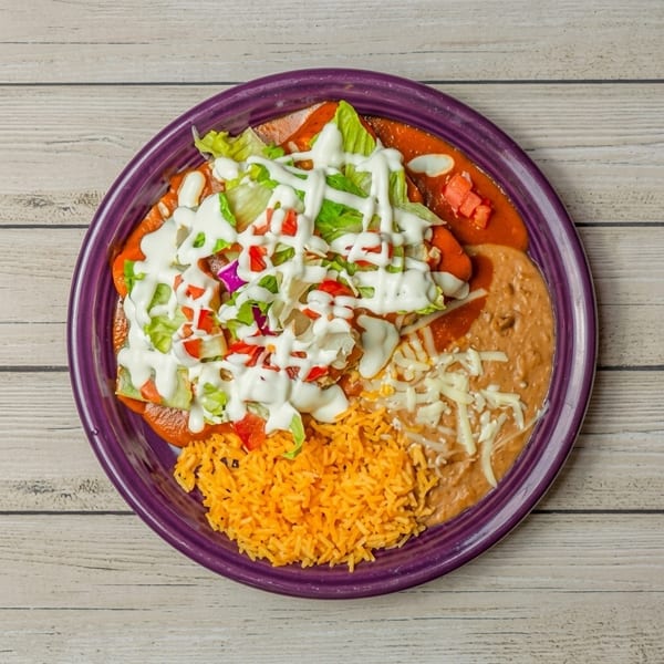 Purple plate with enchiladas topped with lettuce, tomatoes, and sour cream, alongside rice and refried beans.